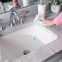 A gray marble bathroom sink with blue toothpaste spilled down the side of the sink basin.  A hand holding a paper towel is wiping up the spill with a paper towel